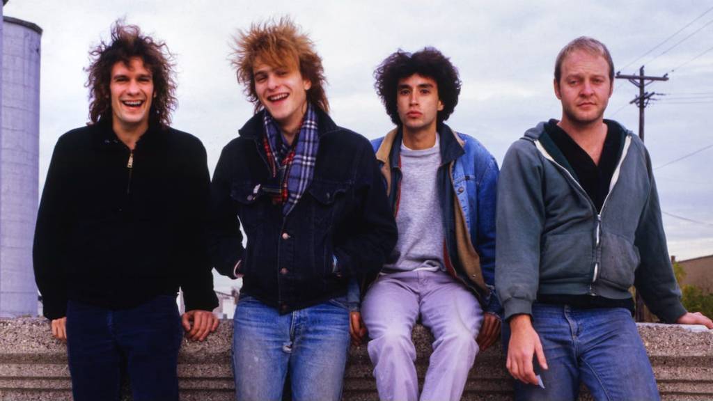 Four young men standing together, smiling in casual clothing, with a cloudy sky in the background.
