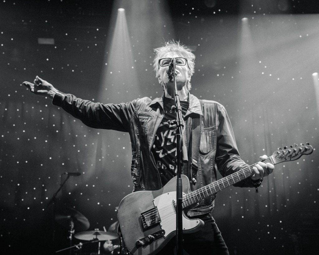 Musical performer on stage with guitar, wearing a leather jacket and glasses, gesturing while singing under stage lights.